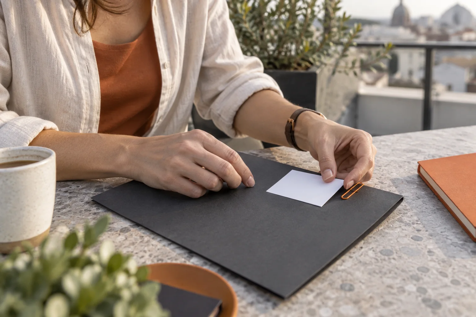 Erwachsene Hände legen eine unbeschriftete Karte auf eine schlichte Mappe an einem Tisch auf einer Dachterrasse.